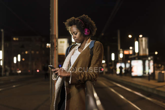 Young woman with headphones and smartphone waiting at tram stop — Stock Photo