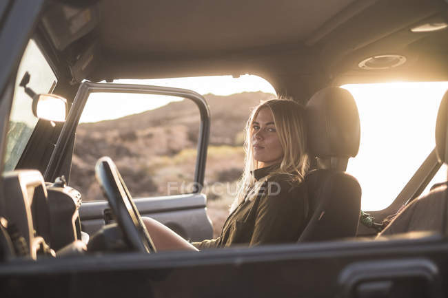 Femme assise en voiture au coucher du soleil et regardant la caméra — Photo de stock