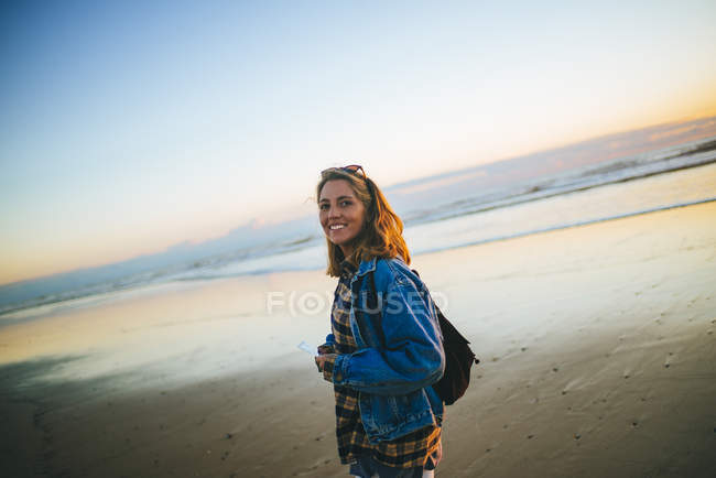 Mujer joven caminando en la playa al atardecer, mirando a la cámara - foto de stock