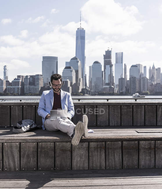 Hombre usando auriculares usando tableta en el paseo marítimo de Nueva Jersey con vista a Manhattan, EE.UU. - foto de stock