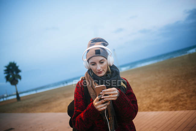 Junge Frau hört Musik auf einem Smartphone, während sie in der Dämmerung am Strand spaziert. — Stockfoto