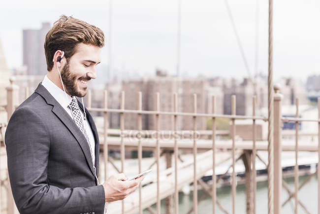 USA, New York, Brooklyn bridge, young businessman with smartphone, cityscape on background — Stock Photo