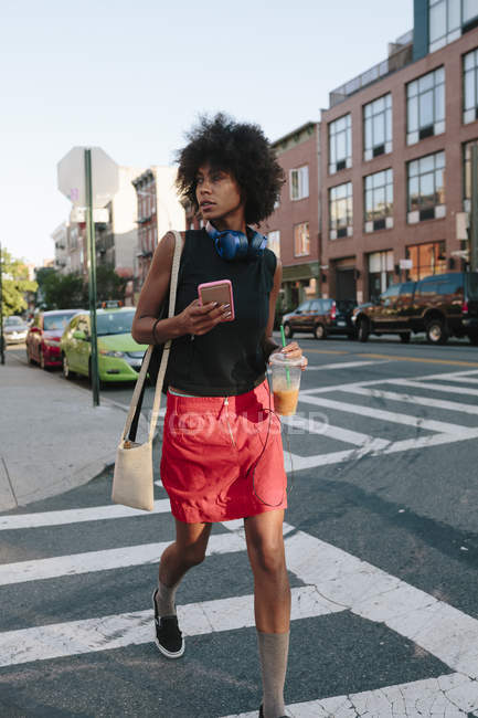 Portrait of young woman walking at street with smartphone and drink — Stock Photo