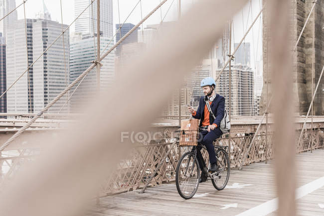 USA, New York, ponte di Brooklyn, Giovane in casco in bicicletta con smartphone, paesaggio urbano sullo sfondo — Foto stock