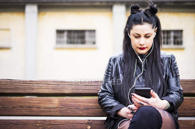 Mujer joven de cabello oscuro sentada en el banco y escuchando música con auriculares y teléfonos inteligentes - foto de stock