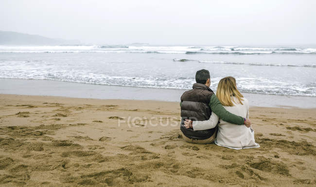 España, Asturias, Vista trasera de pareja abrazándose mirando el mar sentado en la arena de la playa - foto de stock