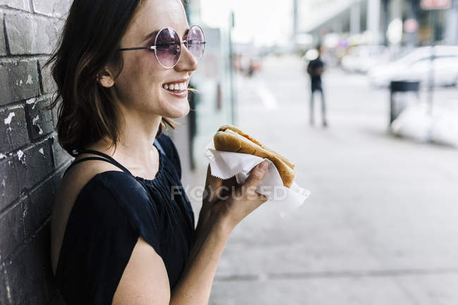 Femme souriante avec Hot Dog appuyé contre le mur — Photo de stock