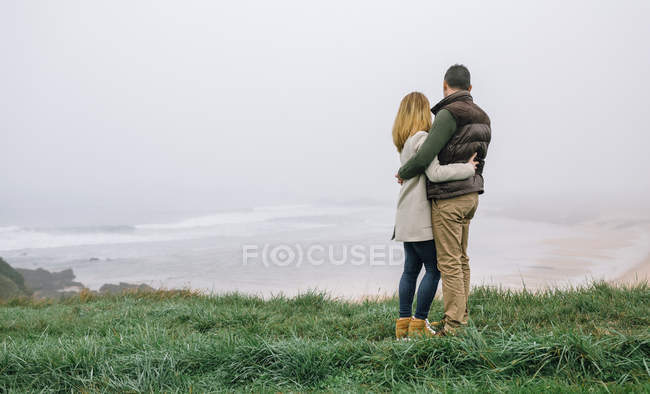 Spagna, Asturie, Indietro vista di coppia irriconoscibile abbracciato guardando la spiaggia in una giornata nebbiosa — Foto stock