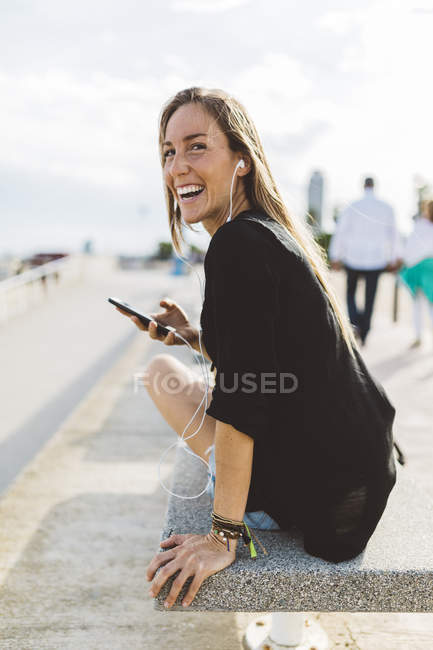 Joyeux jeune femme avec téléphone cellulaire et écouteurs sur la promenade riveraine — Photo de stock