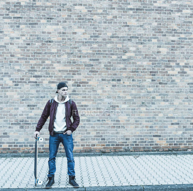 Junger Mann mit Skateboard steht vor Ziegelwand — Stockfoto