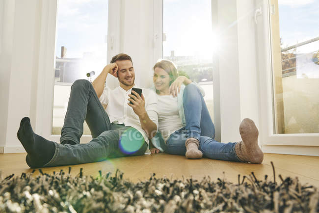 Smiling couple sitting on floor looking at cell phone — Stock Photo