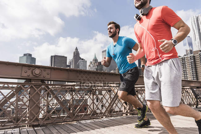 USA, New York, ponte di Brooklyn, due giovani uomini che fanno jogging, vista sul paesaggio urbano sullo sfondo — Foto stock