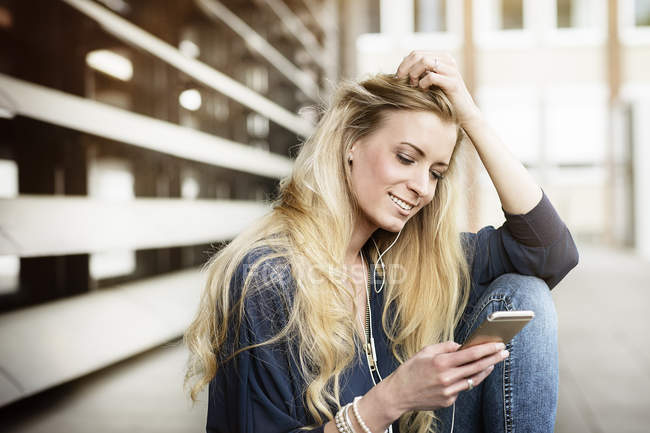 Jeune femme blonde souriante avec écouteurs utilisant un téléphone portable — Photo de stock