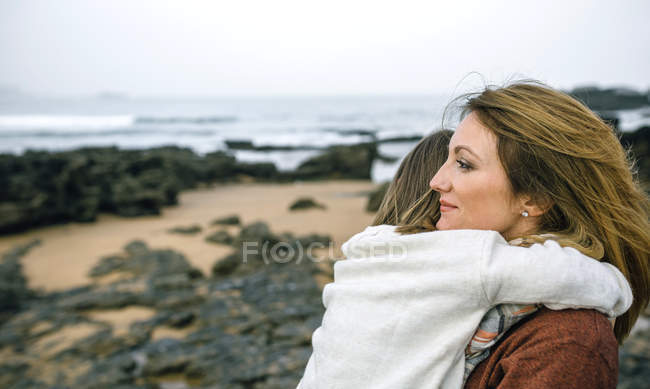 Porträt einer Frau, die beiseite schaut, während sie ein kleines Mädchen am Strand im Winter umarmt, mit dem Meer im Hintergrund — Stockfoto