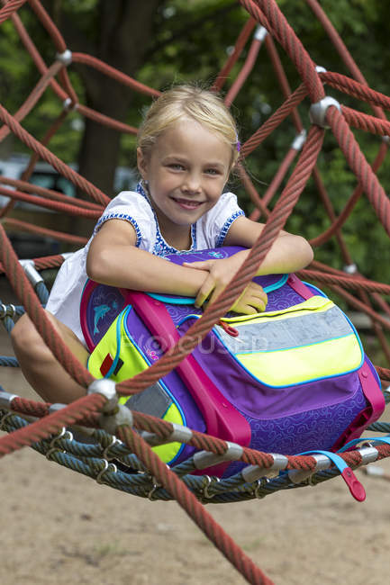 Portrait of smiling little girl with school bag sitting in nest swing on playground — Stock Photo