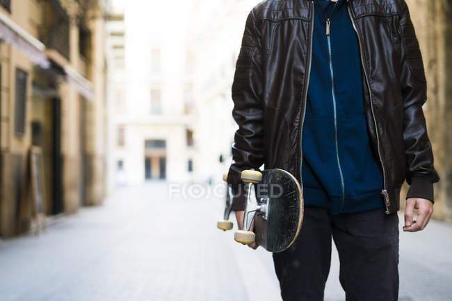 Skater boy sosteniendo monopatín caminando en la calle, vista de sección media - foto de stock