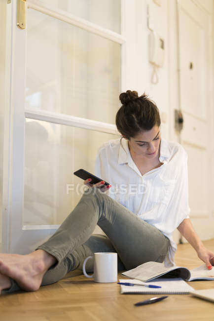 Jeune femme assise sur le sol avec une tasse de café et un smartphone regardant un livret — Photo de stock