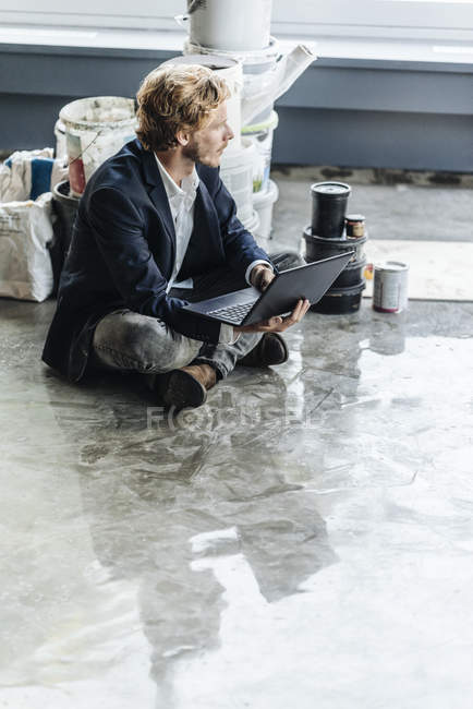 Uomo d'affari seduto sul pavimento con laptop e secchi di vernice sullo sfondo — Foto stock