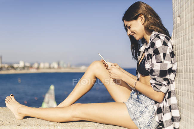 Боковой портрет молодой женщины, сидящей на стене со смартфоном — стоковое фото