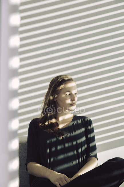Portrait of woman sitting in striped sunshine behind blinds, looking outside — Stock Photo