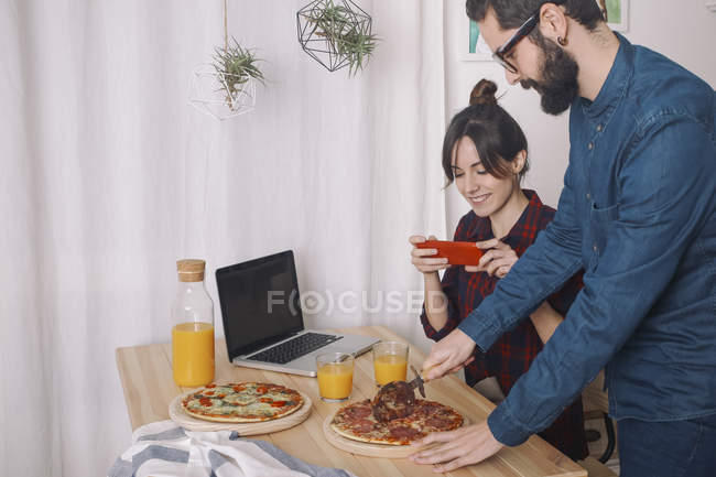 Casal jovem comendo pizza e bebendo suco para o almoço, mulher tirando fotos com smartphone — Fotografia de Stock