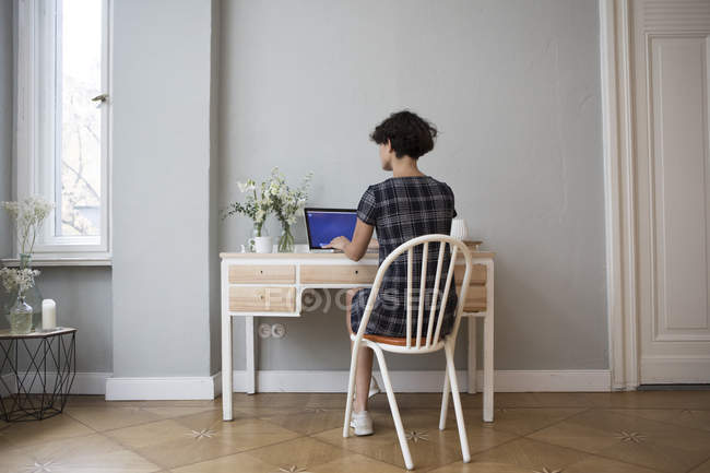 Заднього вигляду молода жінка, сидячи за столом в домашніх умовах і використання ноутбука — стокове фото