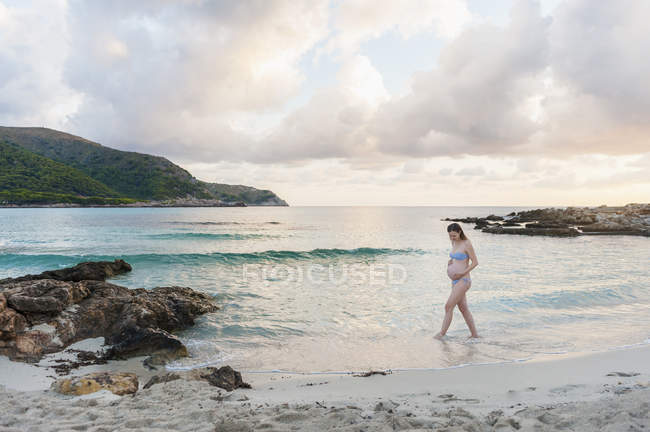 Беременная женщина гуляет по пляжу с облачным небом на заднем плане — стоковое фото