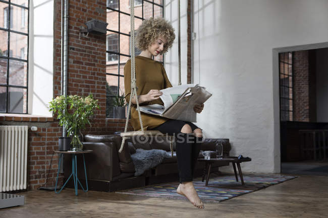 Молода жінка, сидячи на гойдалки у вітальні та читання газет — стокове фото