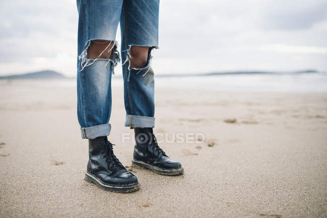 Piernas femeninas con botas y pantalones vaqueros rotos en la playa - foto de stock