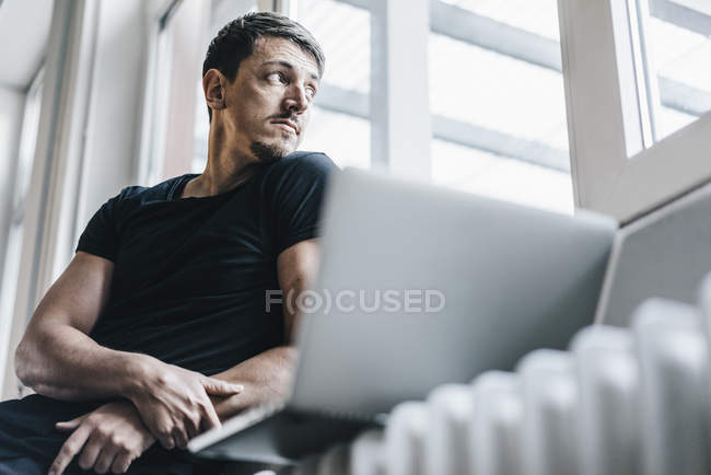 Mann sitzt mit Laptop auf Heizung neben Fenster — Stockfoto