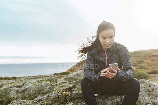 Irlanda, Howth, atleta feminina sentada na costa do penhasco olhando para o telefone celular — Fotografia de Stock