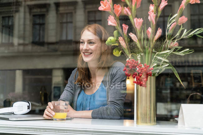 Niedliche rothaarige Kaukasierin sitzt im Café mit Saft im Glas und Blumen in der Vase — Stockfoto