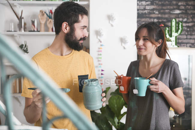 Young woman bringing two cups of tea while boyfriend painting armchair — Stock Photo