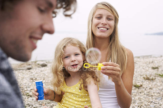 Родители с дочерью на озере пускают мыльные пузыри — стоковое фото
