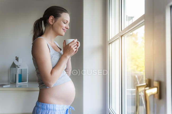 Усміхаючись вагітна жінка тримає Кубок на вікна — стокове фото