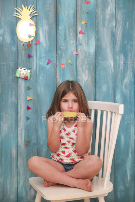 Little girl sitting on chair eating lemon ice lolly — Stock Photo