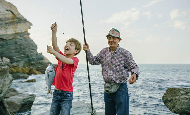 Niño feliz sosteniendo pescado en la línea de pesca capturado por el abuelo - foto de stock