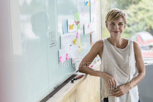 Femme d'affaires souriante debout avec des notes adhésives au tableau blanc dans le bureau moderne — Photo de stock