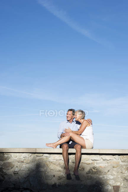 Glückliches Seniorenpaar, das barfuß auf einer Mauer vor dem Himmel sitzt — Stockfoto
