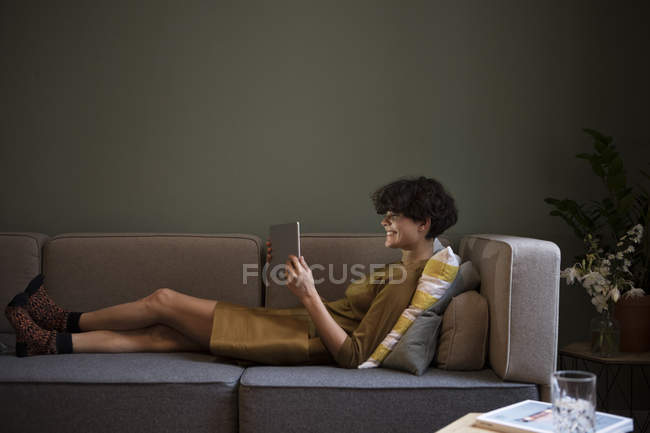 Jeune femme souriante utilisant une tablette sur le canapé à la maison — Photo de stock