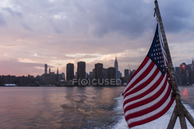 Estados Unidos, Nueva York, horizonte de la ciudad al atardecer y bandera estadounidense en primer plano - foto de stock