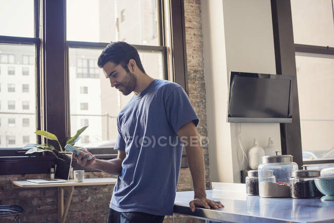Homem com smartphone em pé junto à mesa da cozinha em casa — Fotografia de Stock