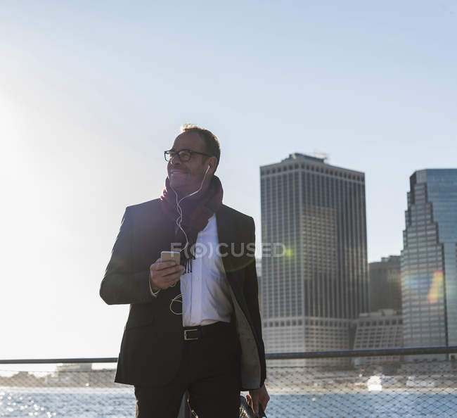 USA, Brooklyn, smiling businessman with earphones and smartphone walking in front of Manhattan skyline — Stock Photo