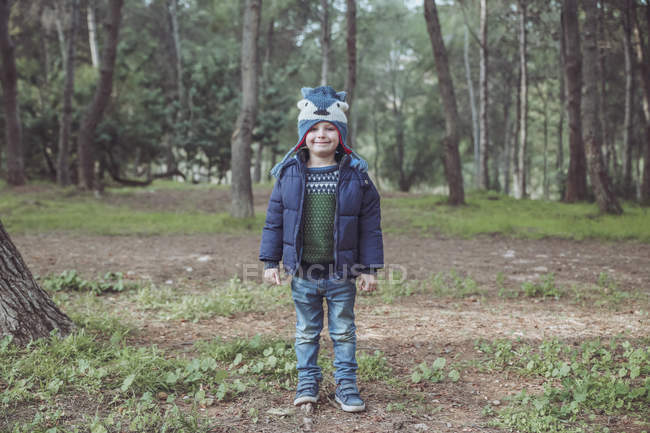 Portrait d'un garçon souriant portant un chapeau laineux debout dans la forêt — Photo de stock
