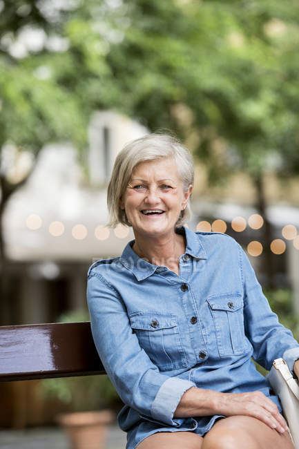 Joyeuse femme adulte âgée assise sur le banc sur fond flou — Photo de stock