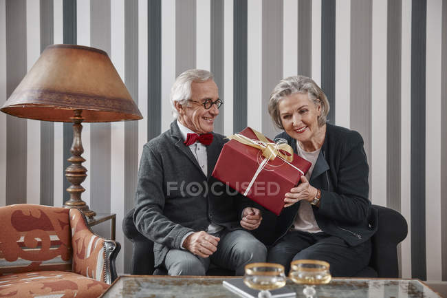 Feliz casal sênior sentado no sofá com presente — Fotografia de Stock