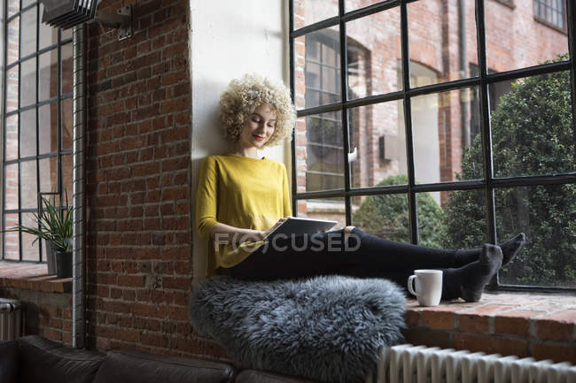 Молода жінка, сидячи на підвіконні і використовуючи цифровий планшетний — стокове фото