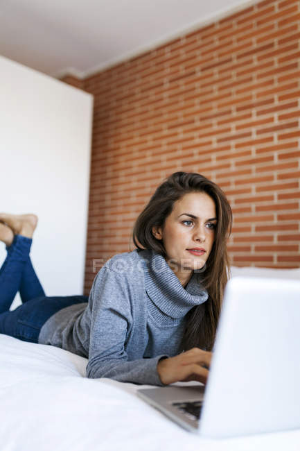 Jeune femme caucasienne attrayante couchée sur le lit et utilisant un ordinateur portable — Photo de stock