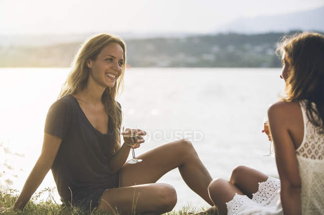 Italy, Lake Garda, two young women sitting at lakeshore with glass of wine — Stock Photo