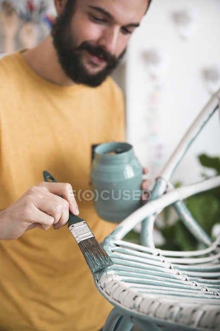 Barbudo hombre pintando sillón de mimbre en casa - foto de stock